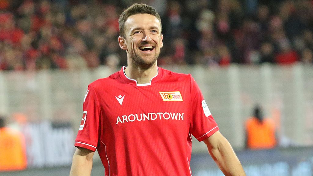 A soccer player in a red jersey beams and looks at the camera, with a cheering crowd in the background.