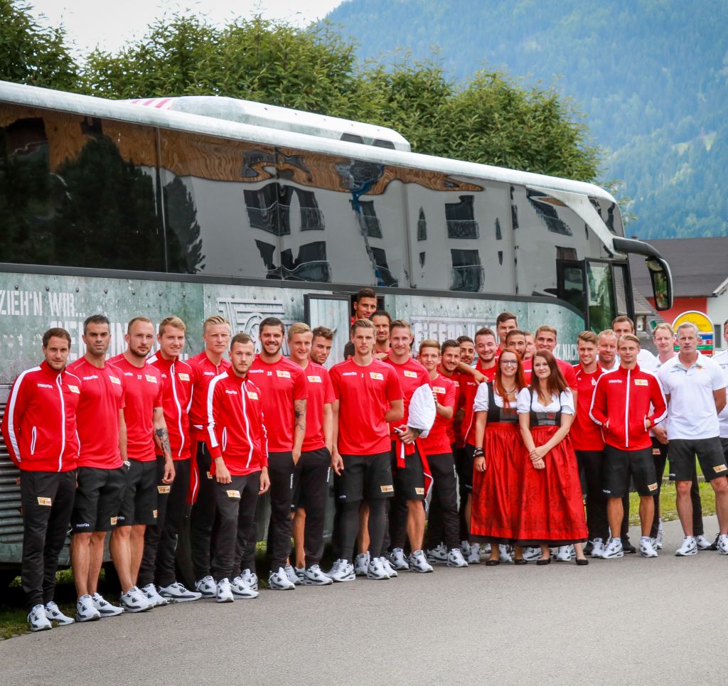 Eine Gruppe von Sportlern in roten Trainingsanzügen posiert vor einem großen Bus, umgeben von Grün und Bergen. Einige tragen traditionelle Trachten.