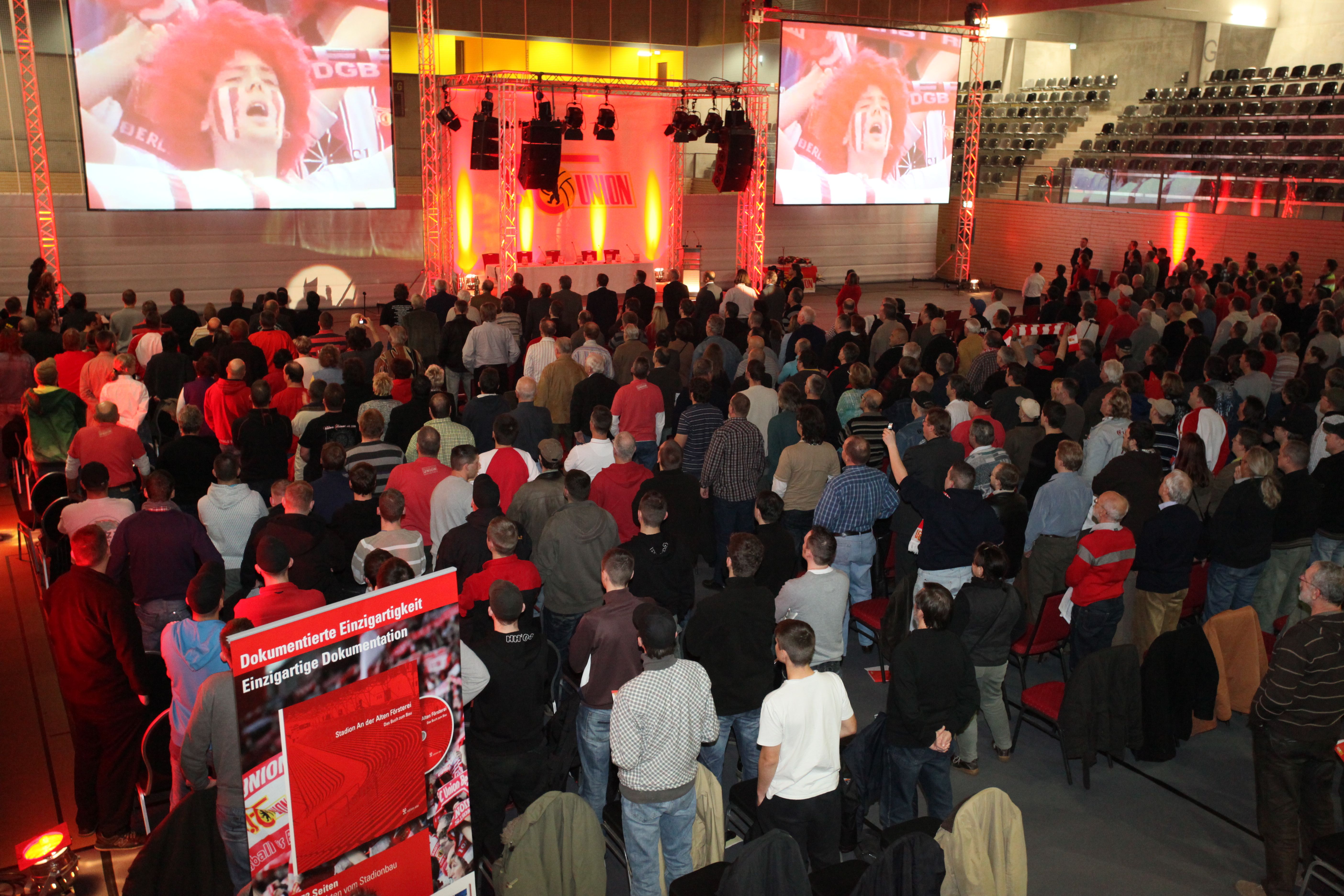 Eine große Menschenmenge steht in einem beleuchteten Veranstaltungsraum, viele Teilnehmer tragen rot-weiße Kleidung und Schals.