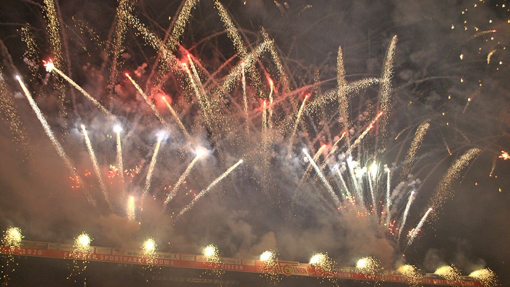Bunte Feuerwerkseffekte erleuchten den Nachthimmel über einem Stadion, umgeben von Rauch und festlicher Stimmung.