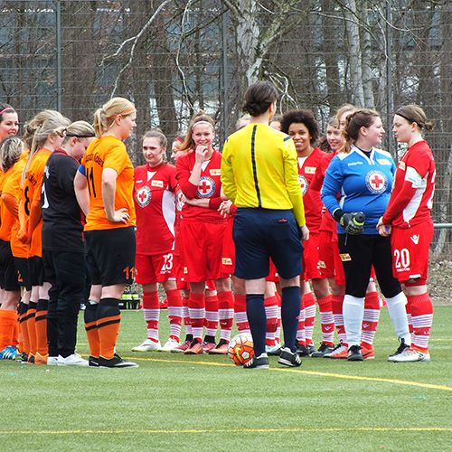 Frauenfußballmannschaften in roten und orangefarbenen Trikots stehen auf dem Feld, während der Schiedsrichter die Anweisungen gibt.