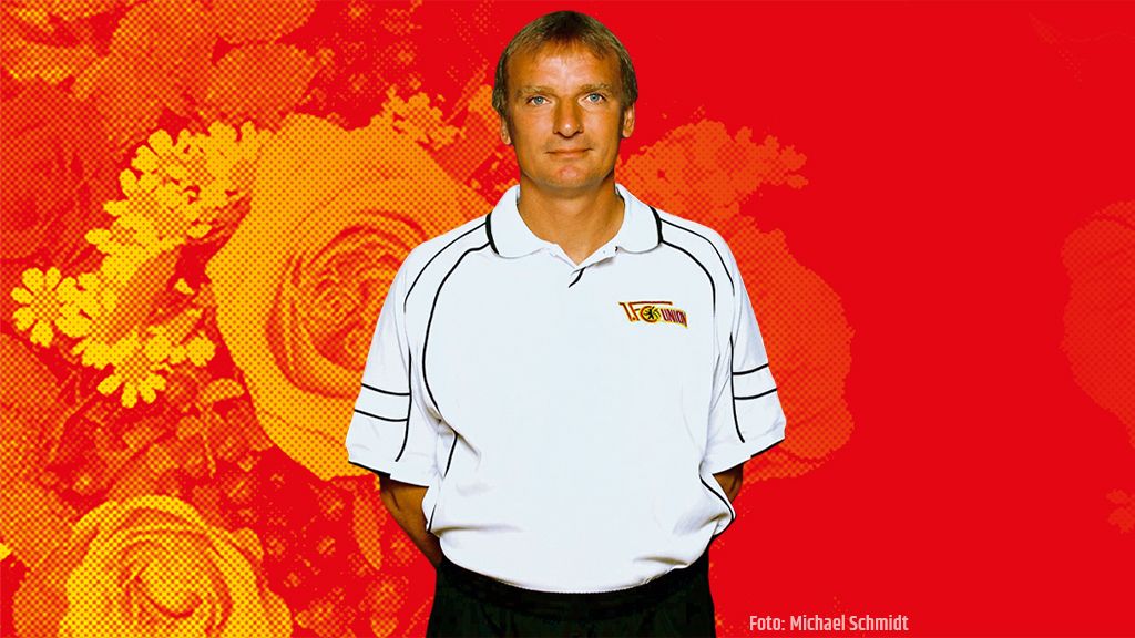 Ein Mann in einem weißen Polo-Shirt mit Logo steht vor einem rot-gelben, floral gemusterten Hintergrund.
