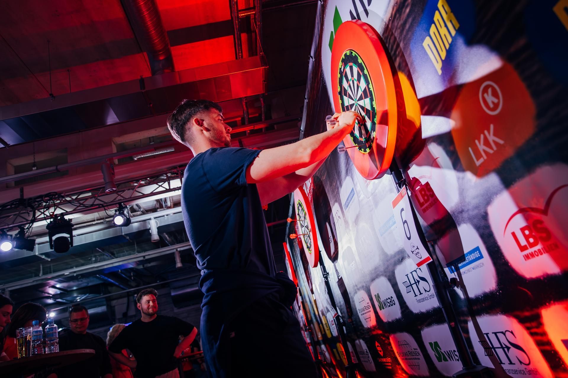 Ein Dartspieler zielt auf eine Dartscheibe bei einem Turnier, während Zuschauer im Hintergrund stehen. Intensives Licht beleuchtet die Szene.