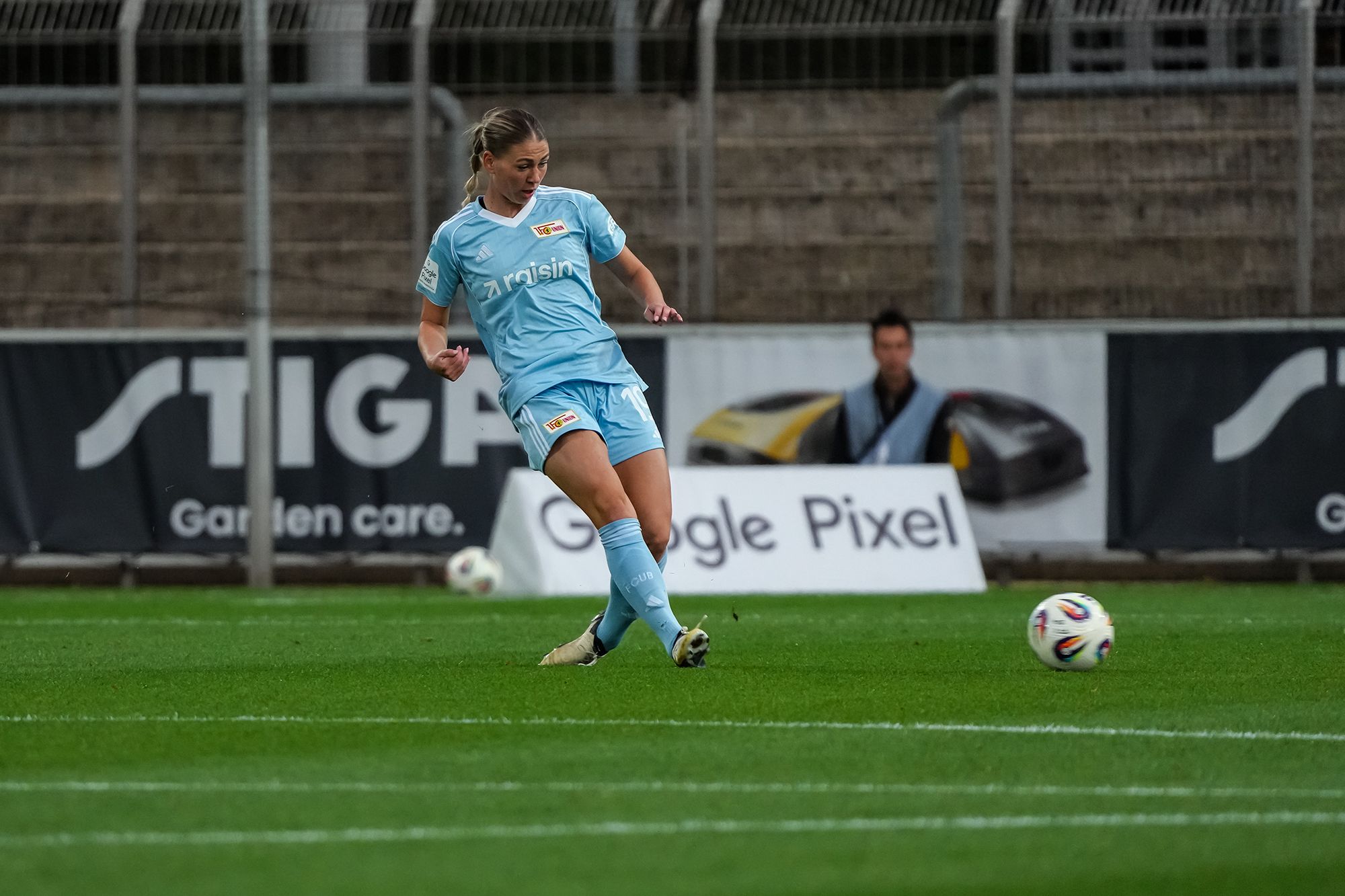 Eine Fußballspielerin in blauer Uniform tritt einen Ball auf einem Rasenplatz. im Hintergrund sind Zuschauer und Werbebanner zu sehen.