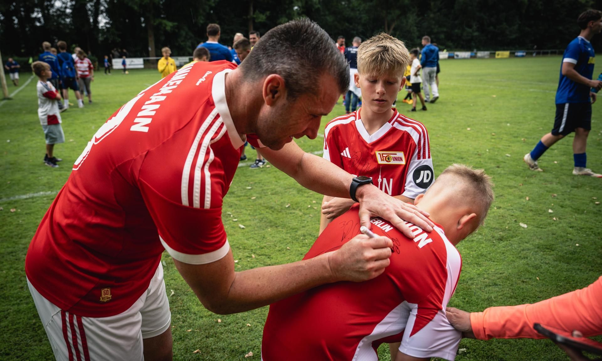 Ein Trainer signiert das Trikot eines jungen Spielers auf einem Fußballplatz, während andere Kinder im Hintergrund spielen.