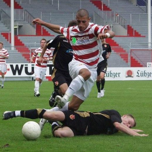 Ein Fußballspieler springt über einen am Boden liegenden Gegner, während ein weiteres Spiel im Hintergrund stattfindet.