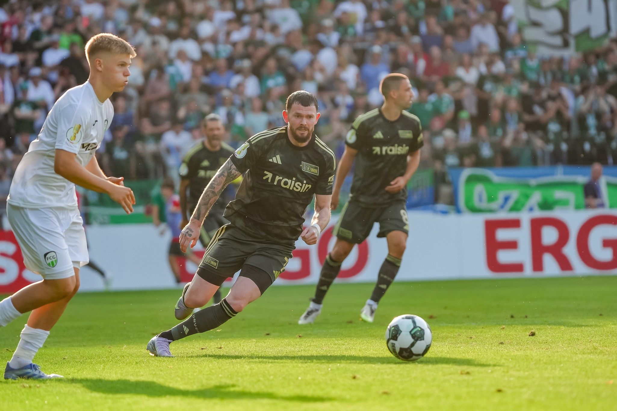 Spielszene im Fußball mit zwei Spielern in Aktion und Zuschauern im Hintergrund. Ein Spieler führt den Ball.