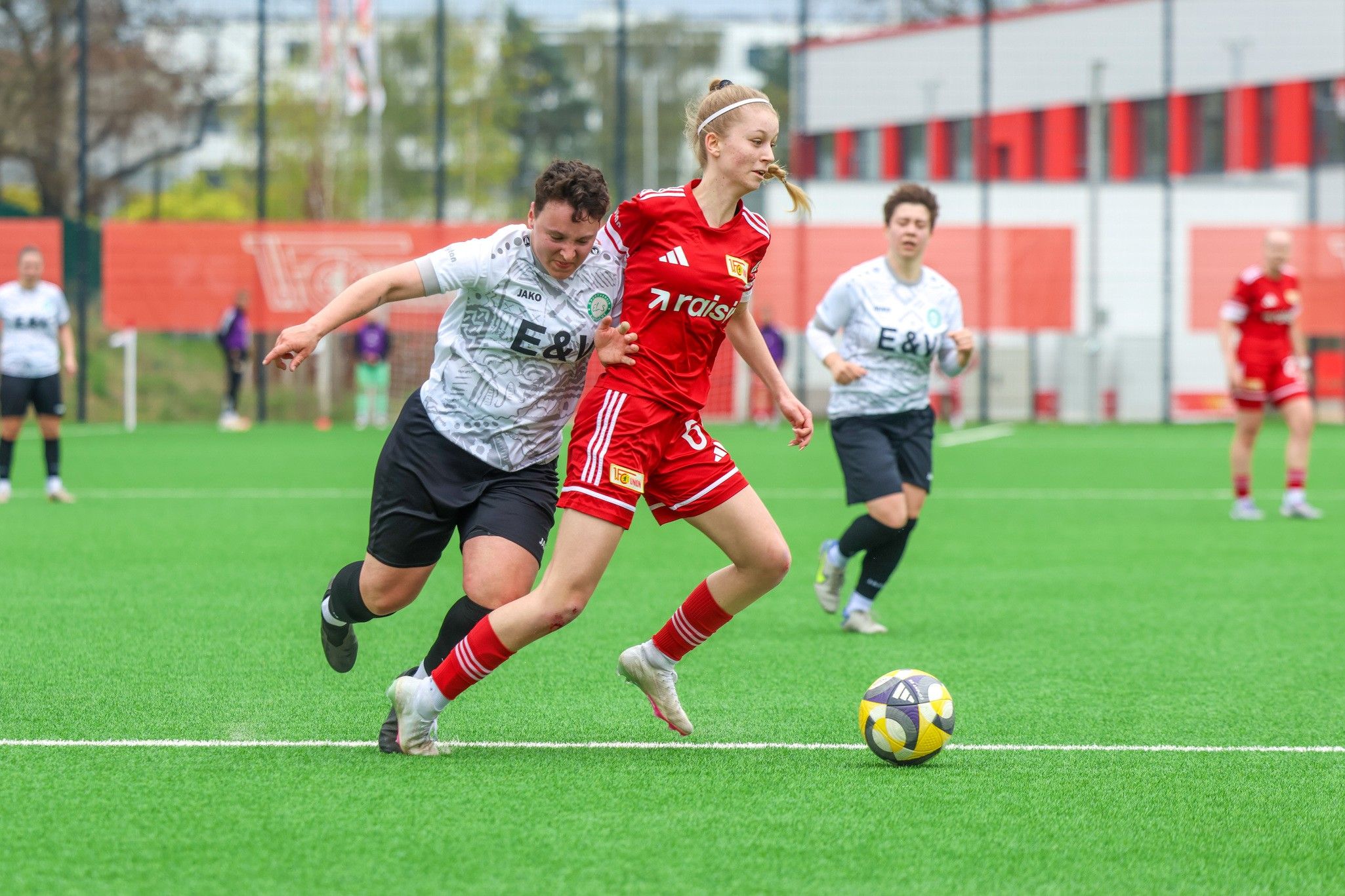 Eine Fußballspielszene mit zwei Spielerinnen, eine in rotem Trikot und die andere in einem grauen Trikot, die um den Ball kämpfen.