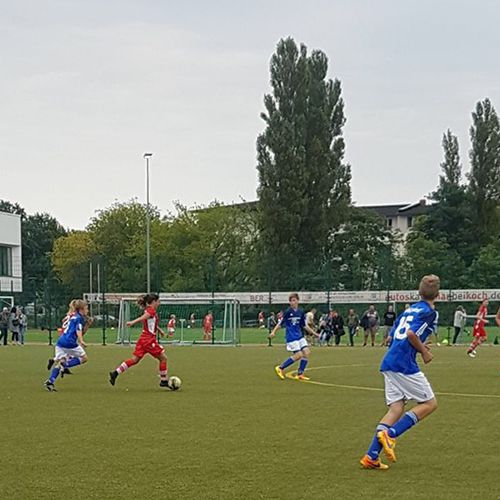 Jugendfußballspiel auf einem Kunstrasenplatz mit Spielern in roten und blauen Trikots. Hintergrund zeigt Bäume und Gebäude.
