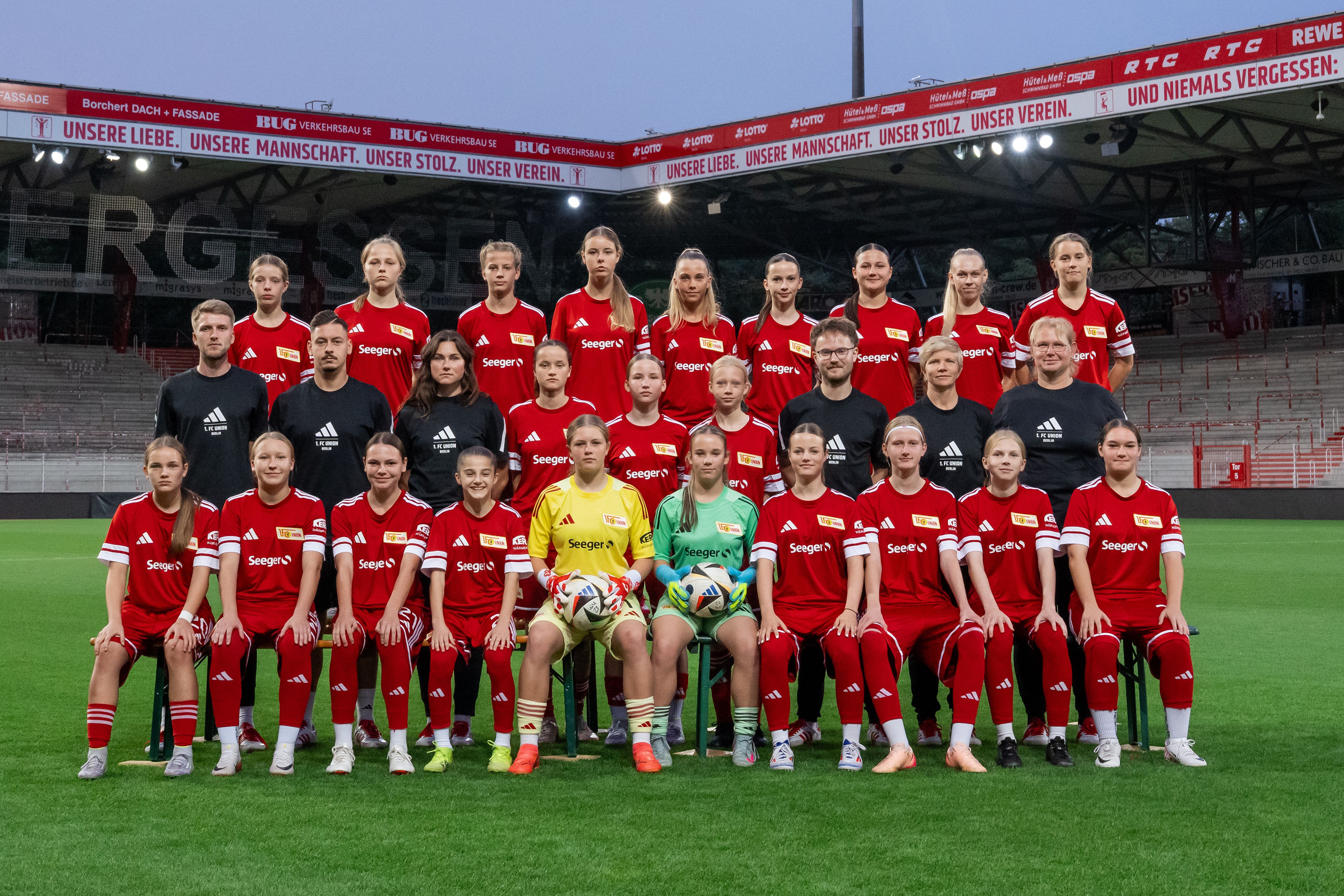Ein Fußballteam in roten Trikots posiert auf dem Rasen eines Stadions, umgeben von leeren Tribünen.