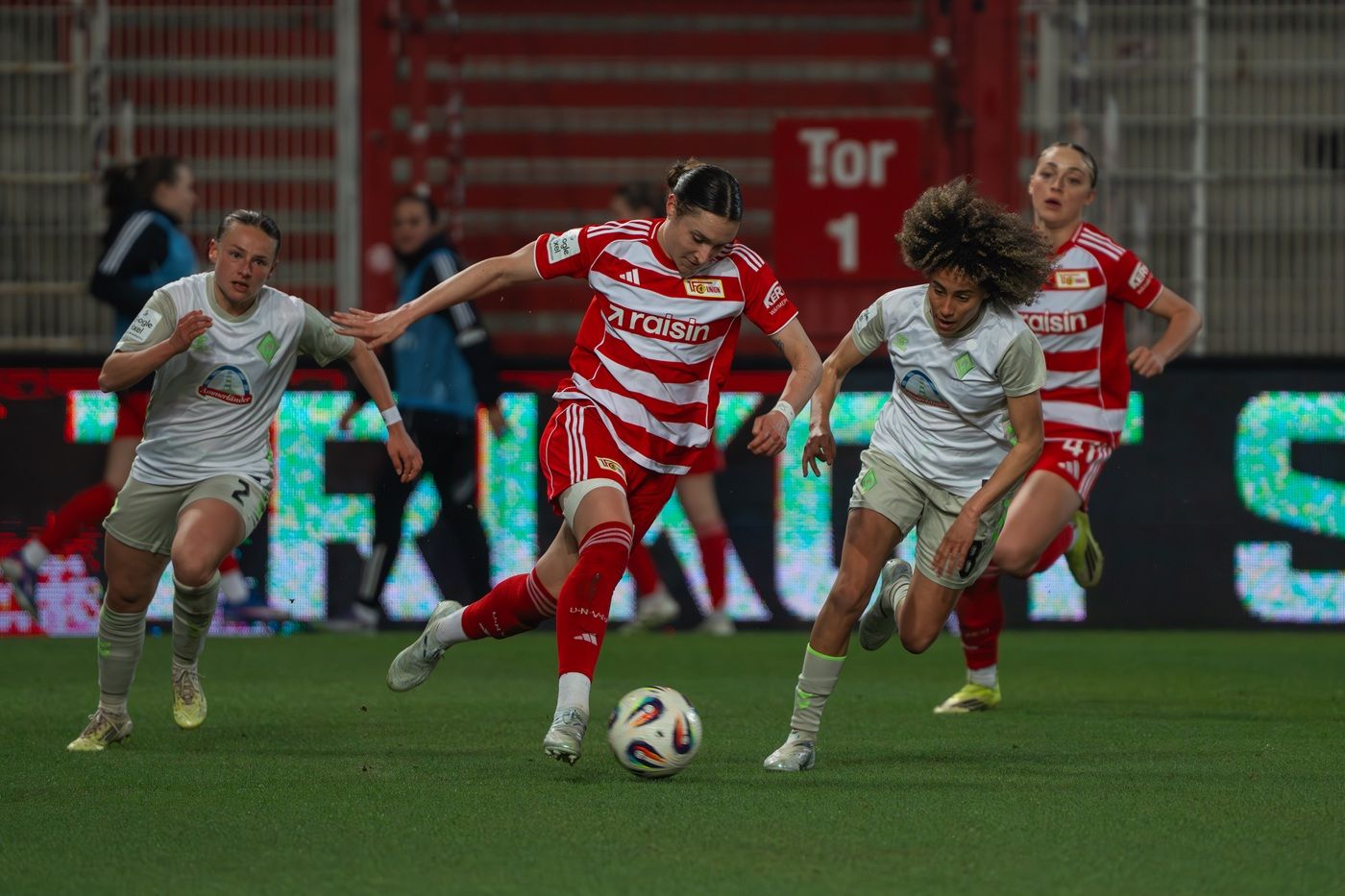 Frauenfußballspiel: Spielerinnen kämpfen um den Ball auf einem grünen Spielfeld vor einer LED-Wand.