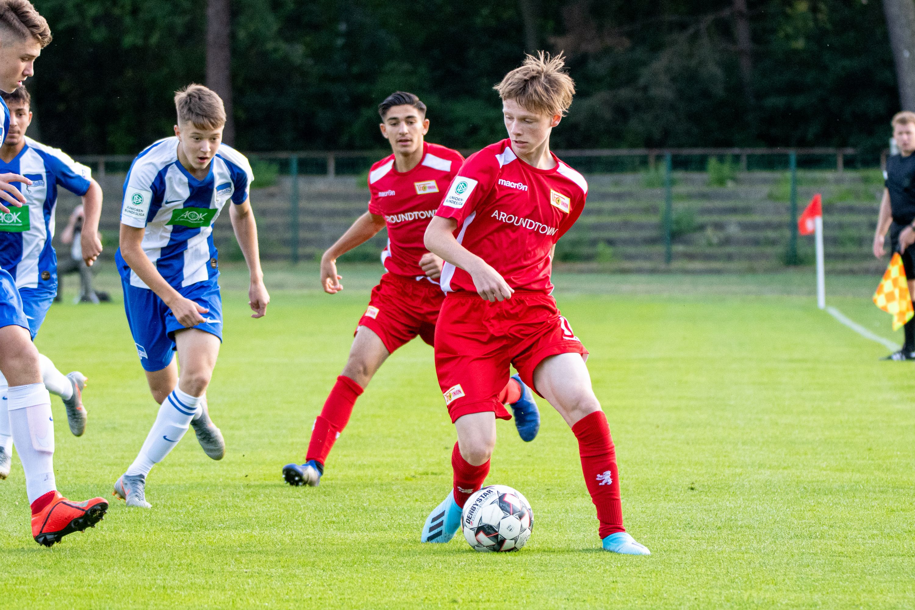 Ein Jugendfußballspiel mit Spielern in roten und blauen Trikots auf einem grünen Rasenplatz.