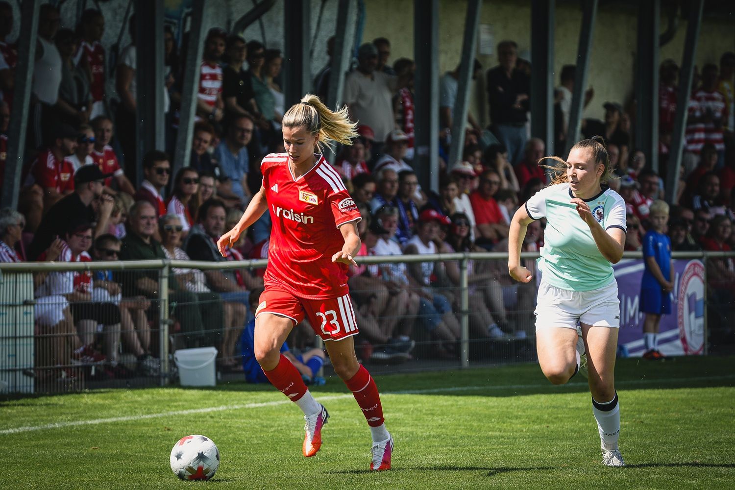 Eine Fußballspielerin in rotem Trikot dribbelt den Ball, während eine Gegnerin in grünem Trikot verfolgt. Zuschauer im Hintergrund.