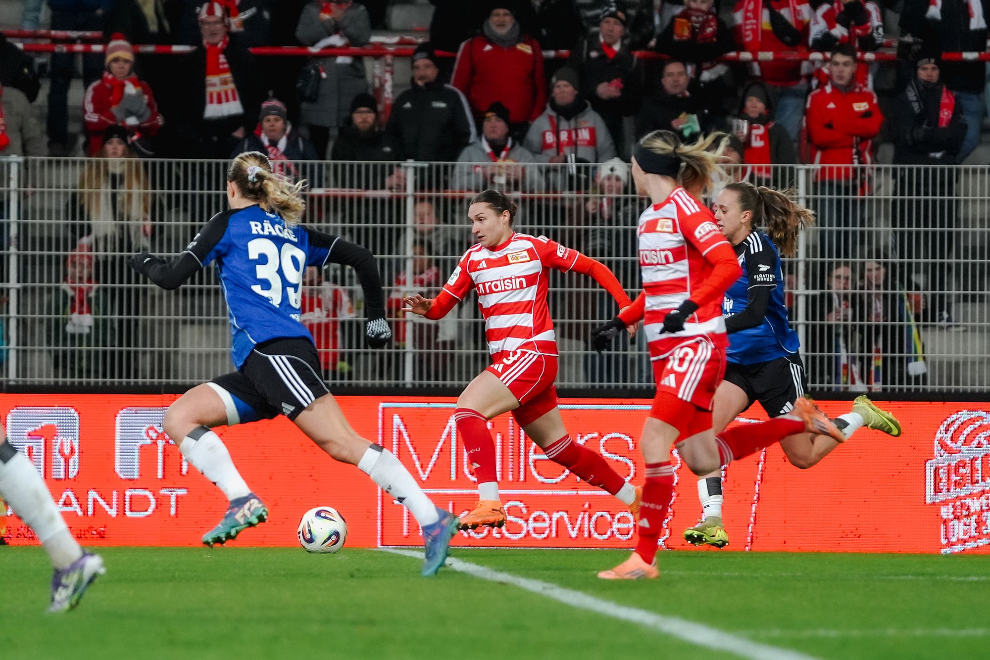 Eine Fußballspielszene mit Spielerinnen in roten und weißen Trikots, die auf ein Tor zulaufen, während eine Torhüterin sich positioniert.