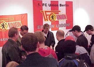Eine Gruppe von Menschen steht vor einer Wand mit dem Banner des 1. FC Union Berlin und diskutiert.