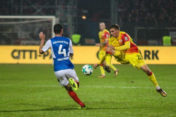 A soccer player jumps into the air to play the ball while another player chases him.