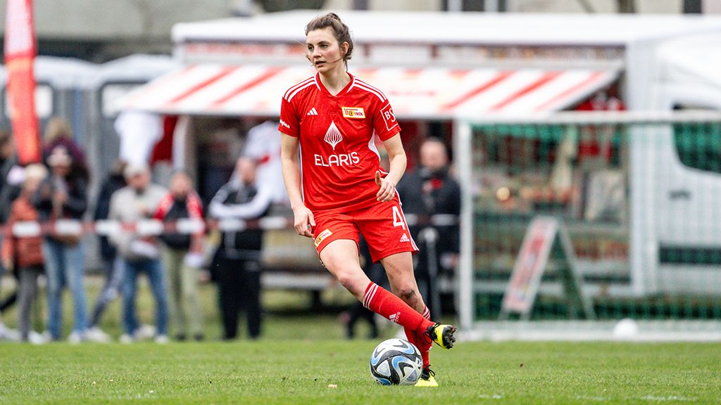 Eine Fußballspielerin in rotem Trikot dribbelt einen Ball auf einem Rasenfeld. Zuschauer sind im Hintergrund zu sehen.