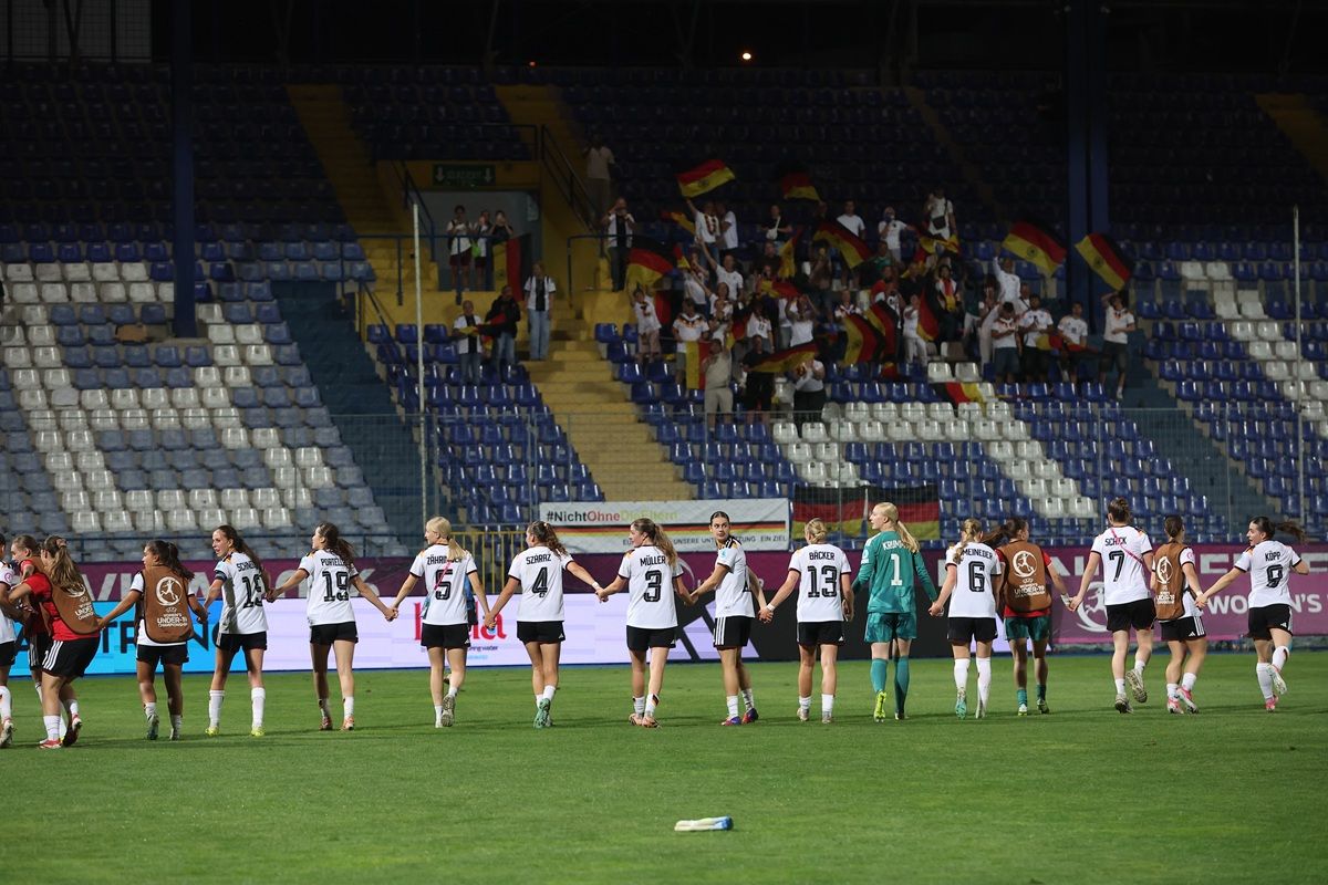 Frauenfußballmannschaft steht am Spielfeldrand, während Fans mit Flaggen im Hintergrund jubeln.