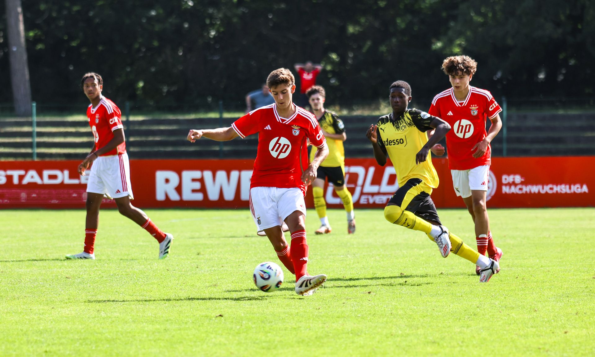 Spielszene eines Fußballspiels mit Spielern in roten und schwarzen Trikots auf einem grünen Rasenplatz.