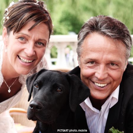 Braut und Bräutigam lächeln fröhlich vor der Kamera, während ein schwarzer Hund zwischen ihnen posiert.