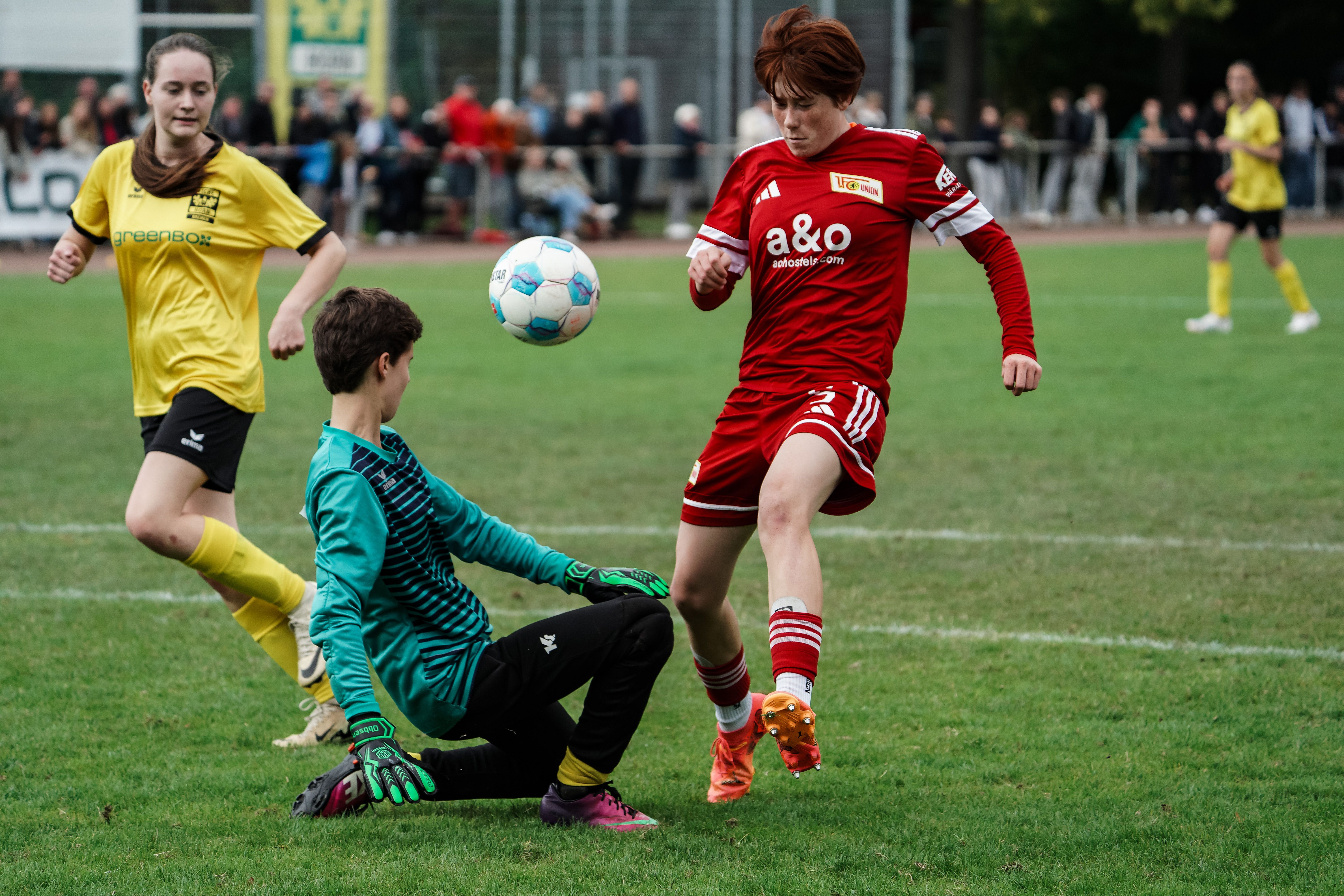 A soccer game between two teams, a player in red jerseys dribbles the ball while a goalkeeper in green avoids it.