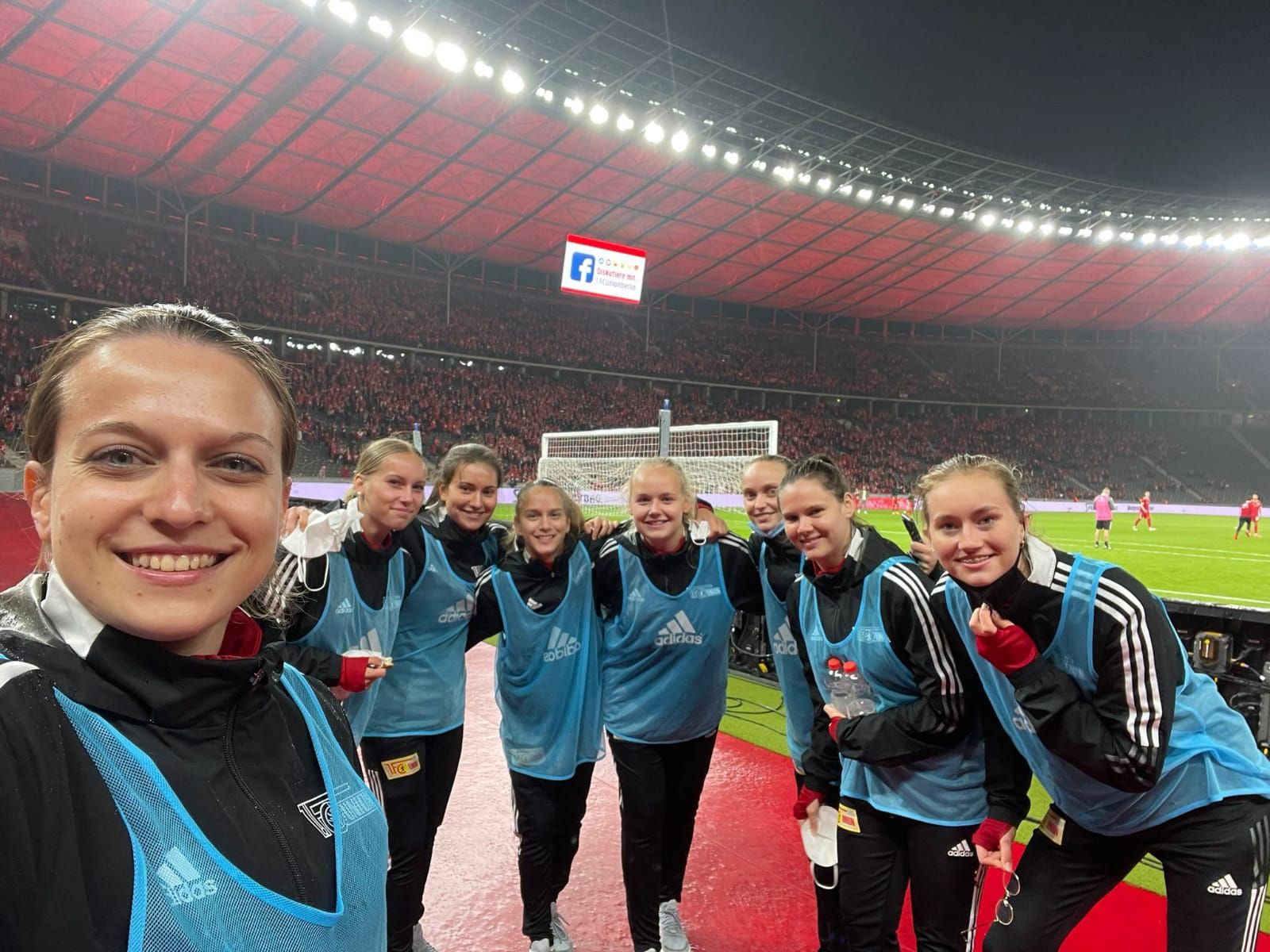Junge Frauen in Sportkleidung lächeln für ein Selfie vor einem Fußballfeld mit Zuschauern im Hintergrund.