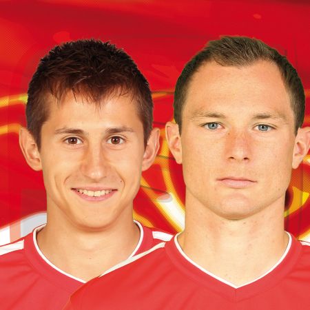 Zwei Fußballspieler in roten Trikots vor einem roten Hintergrund, einer lächelt, der andere schaut ernst.