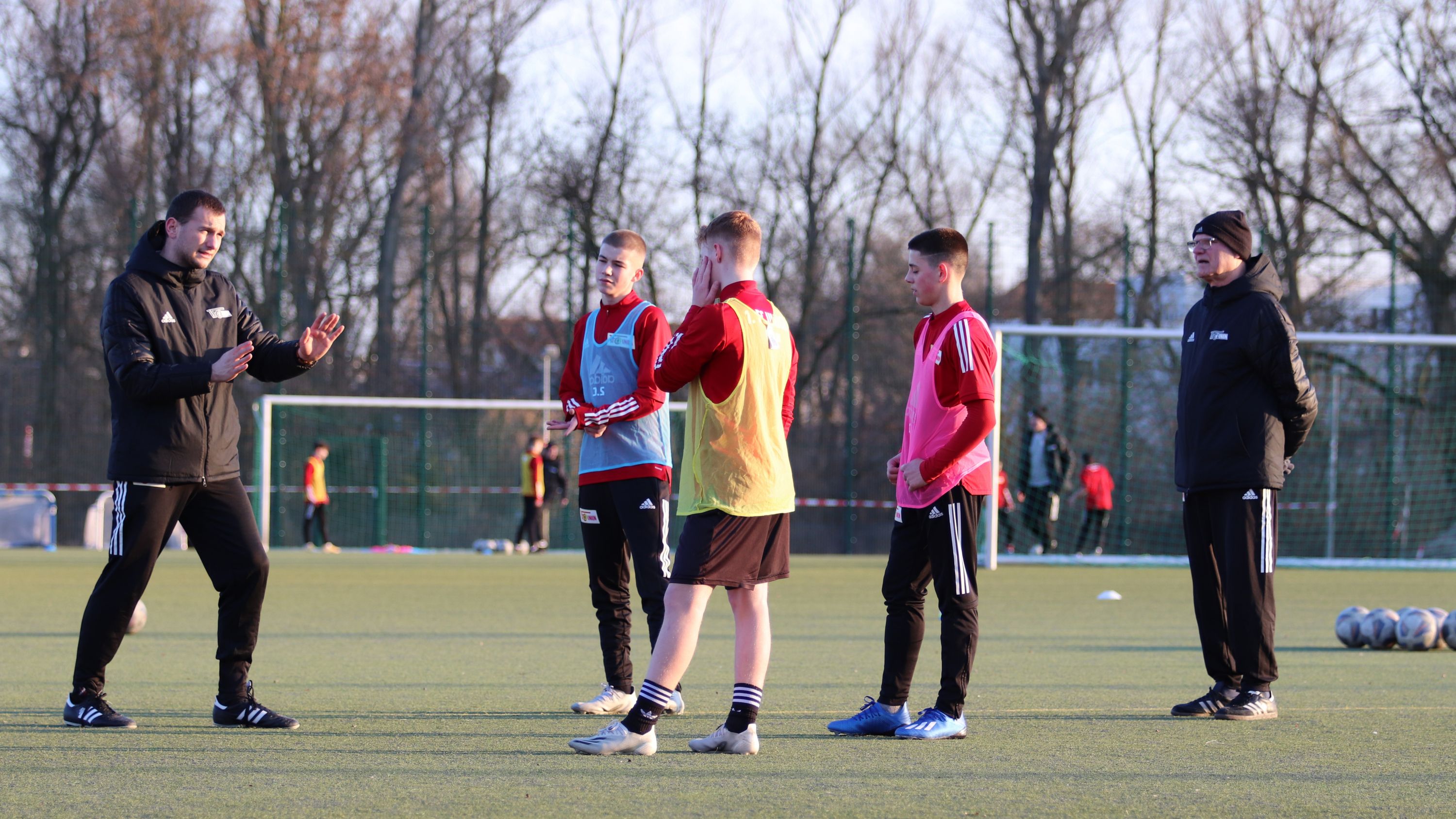 Fünf Fußballspieler in Trainingskleidung stehen auf einem Platz und hören einem Trainer zu, während andere im Hintergrund trainieren.