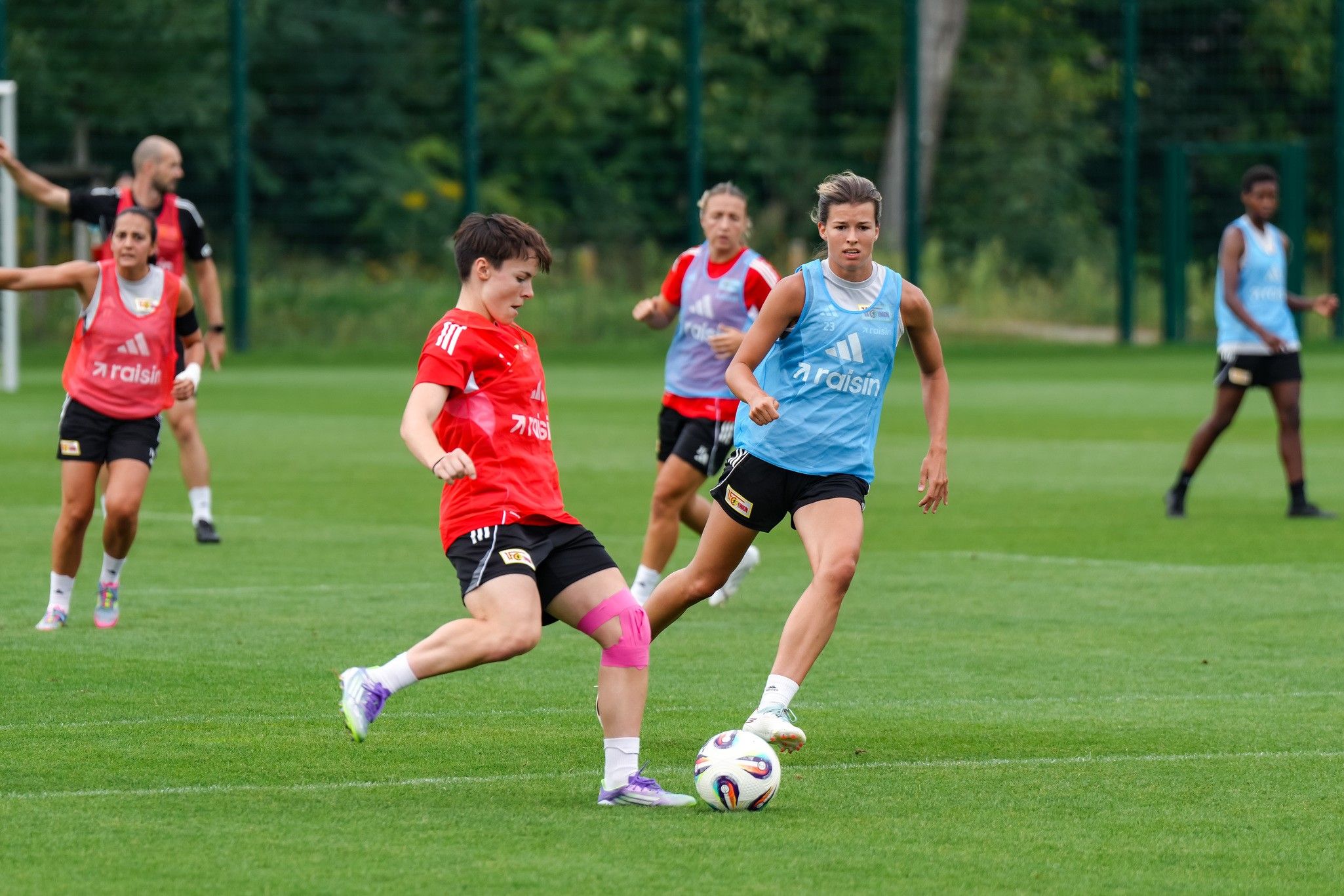 Zwei weibliche Fußballspielerinnen in Trainingsoutfits spielen auf einem grünen Fußballplatz.