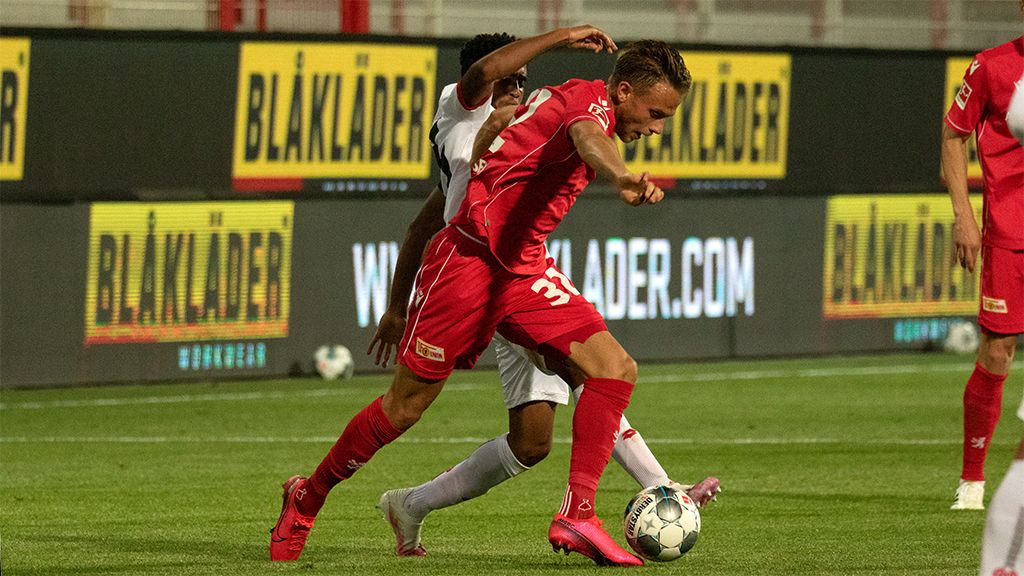 Ein Fußballspieler trägt ein rotes Trikot. Er kämpft um den Ball. Ein Gegner ist bei ihm. Im Hintergrund sind Werbebanner.