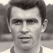 Schwarz-weiß Porträt eines männlichen Fußballspielers mit kurzem Haar und ernstem Ausdruck, im Hintergrund unscharfe Landschaft.