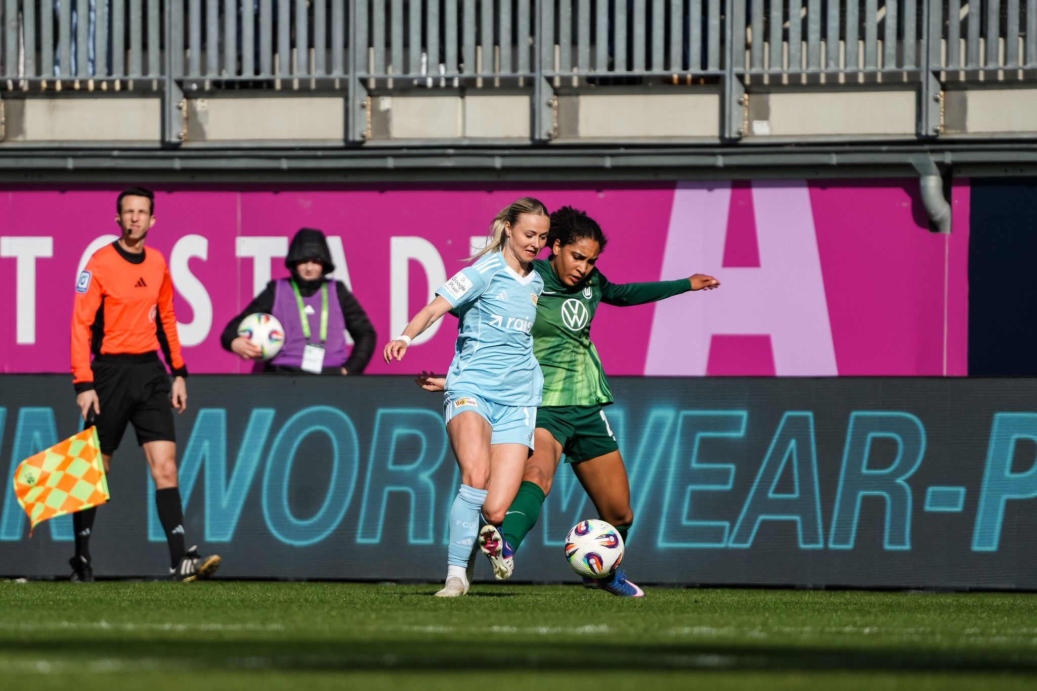 Zwei Fußballspielerinnen kämpfen um den Ball, während ein Schiedsrichter und ein Linienrichter am Spielfeldrand stehen.