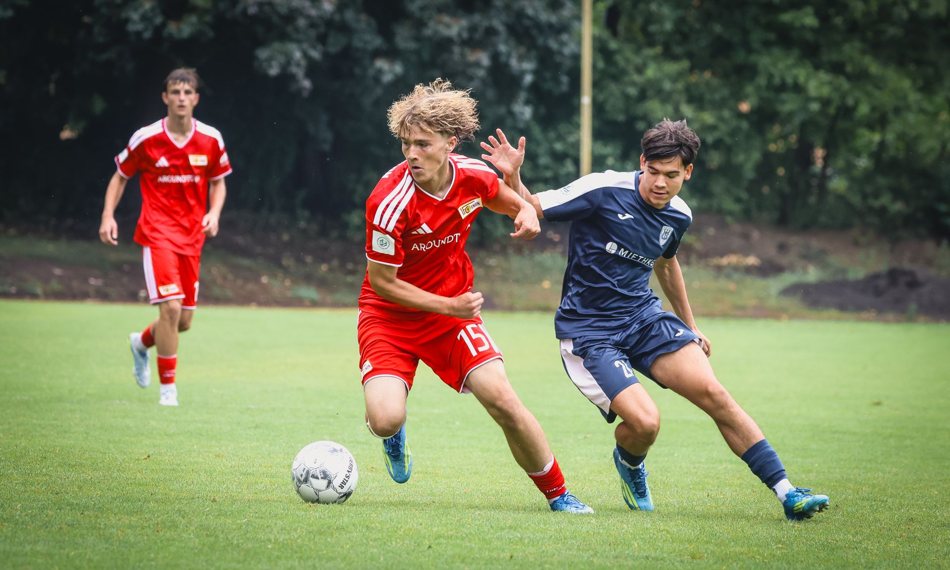 Zwei Fußballspieler in actionreicher Spielsituation, ein Spieler in rotem Trikot dribbelt, während der andere in blauem Trikot versucht, ihn zu stoppen.