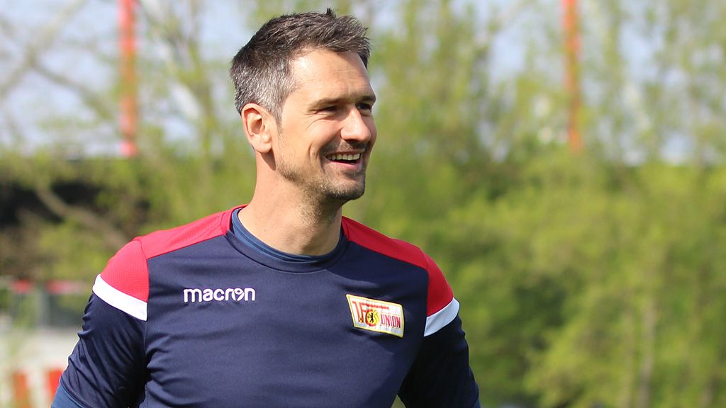 A smiling man in sportswear stands outside in front of a green background.