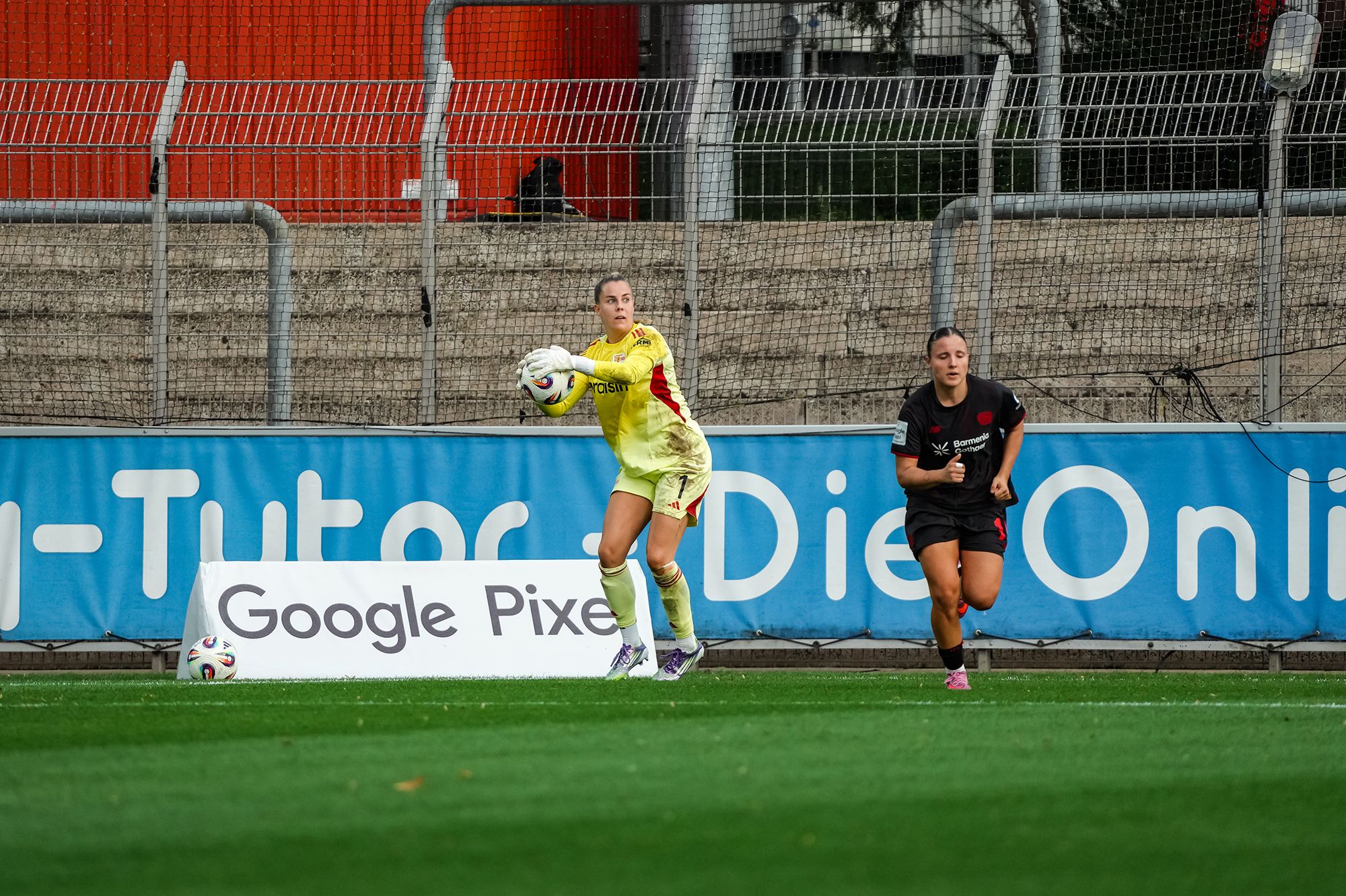 Eine Torhüterin in gelben Trikots fängt den Ball, während eine Spielerin in Schwarz im Hintergrund läuft.