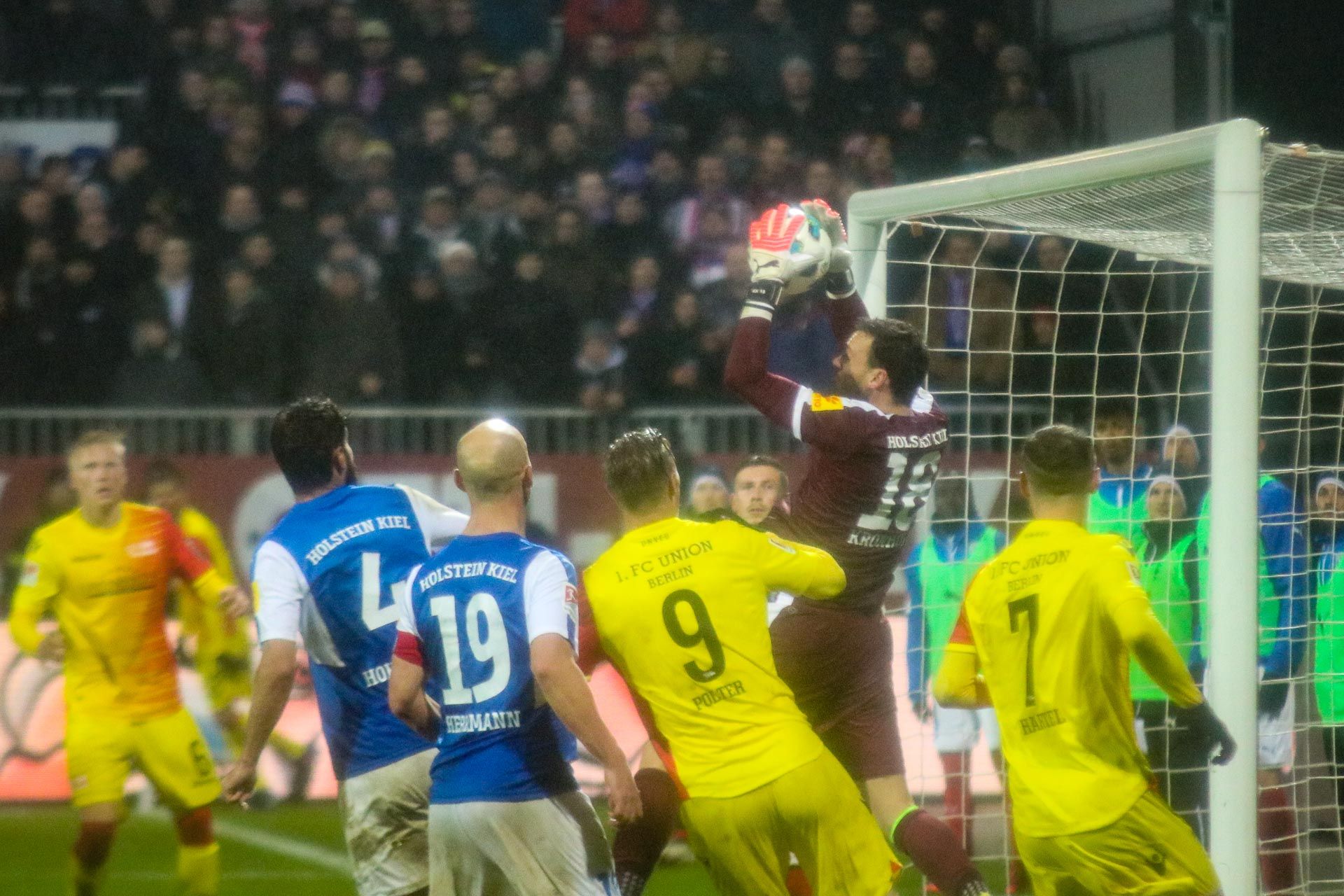 Ein Torwart springt. Er will einen Schuss halten. Es ist ein Fußballspiel. Viele Spieler sind da. Sie tragen verschiedene Trikots.