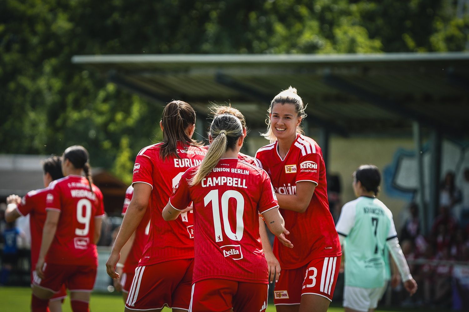 Vier Fußballspielerinnen in roten Trikots feiern auf dem Spielfeld. Im Hintergrund ist eine Sporteinrichtung und Zuschauer zu sehen.