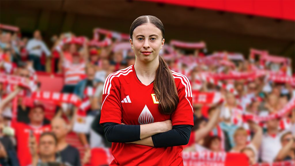 A young woman in a red football jersey stands with her arms crossed in front of a cheering crowd.