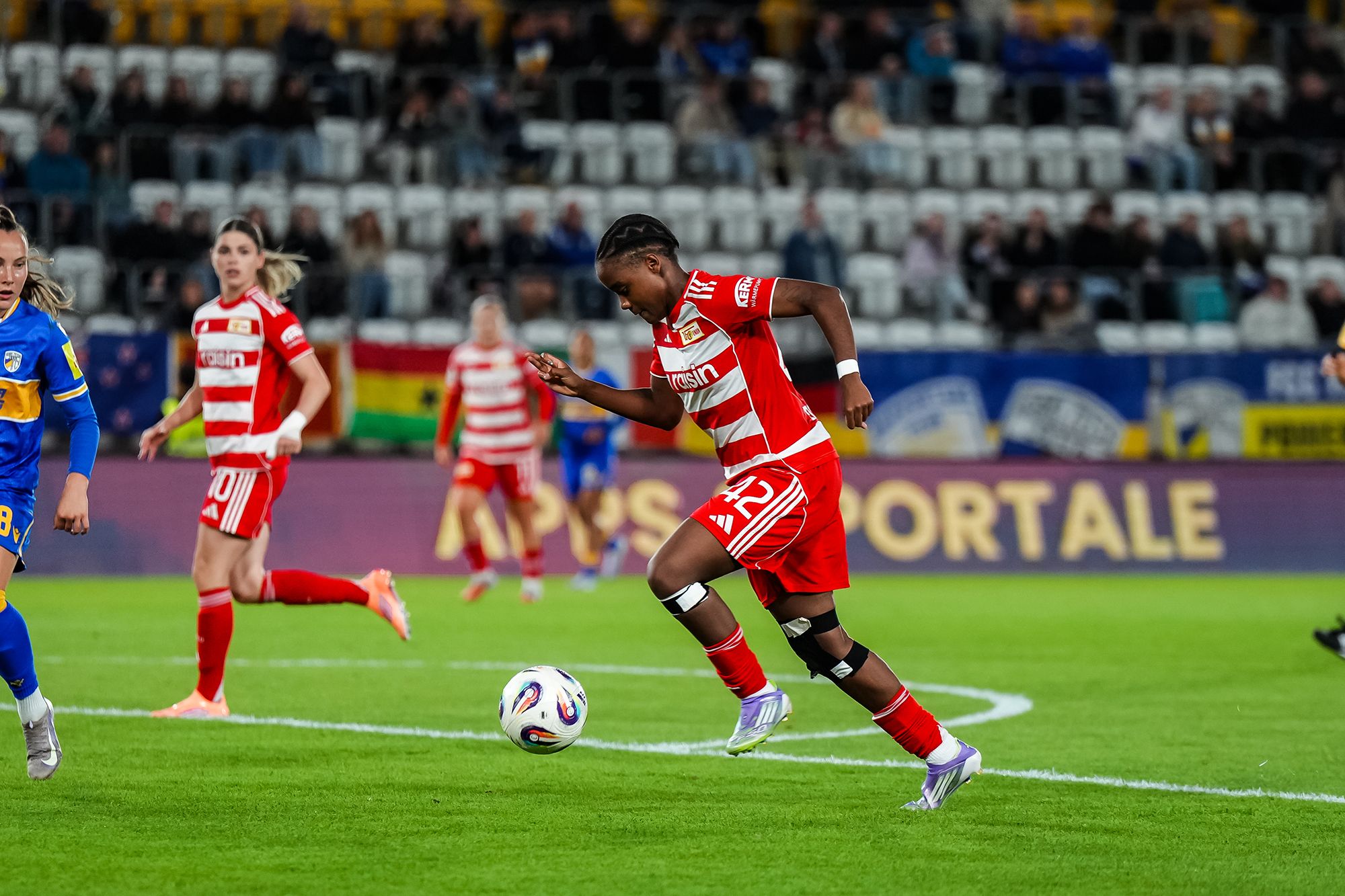 Eine Spielerin in rotem Trikot dribbelt den Ball während eines Fußballspiels auf dem Rasen. Im Hintergrund sind weitere Spielerinnen zu sehen.