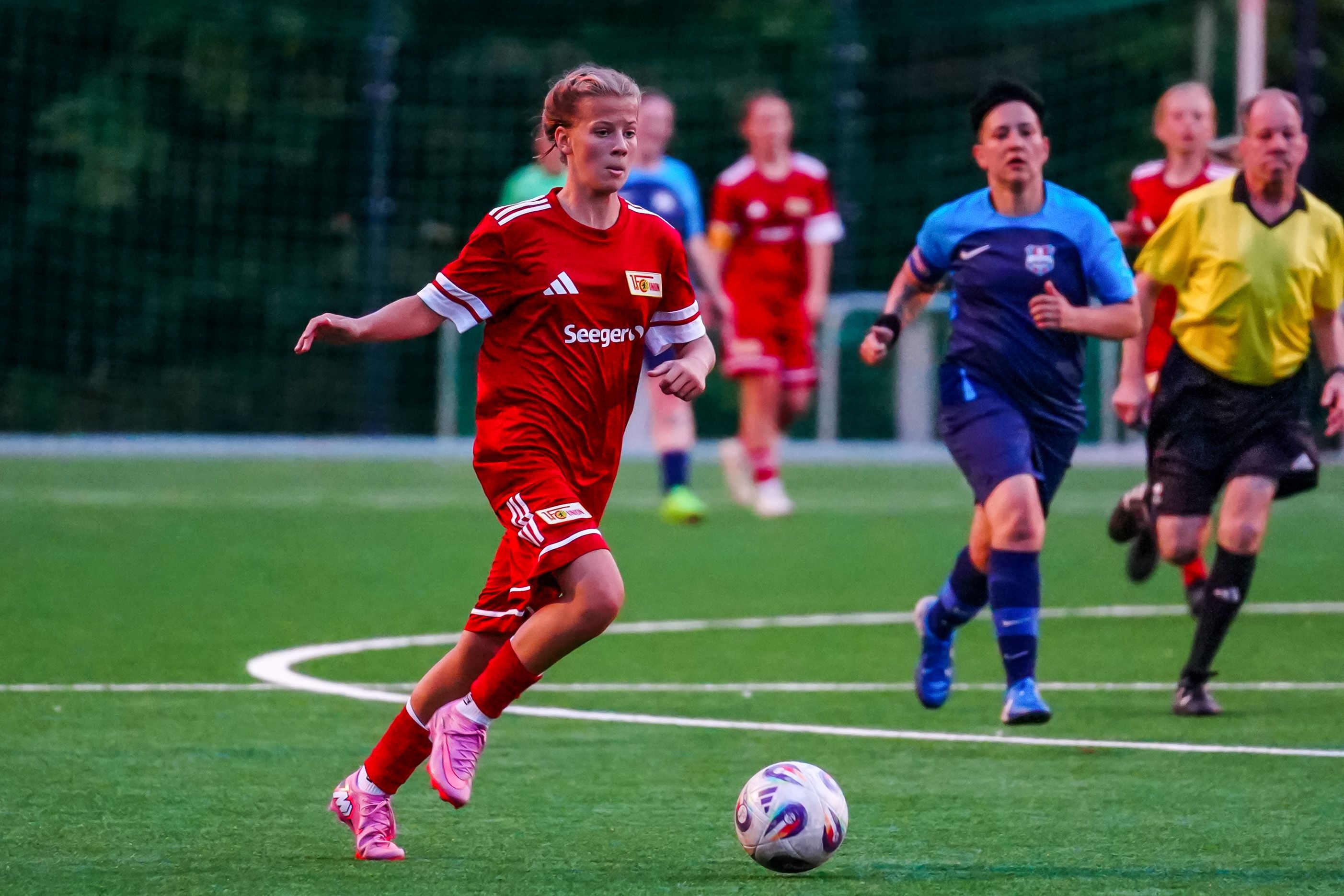 Ein Mädchen trägt ein rotes Fußballtrikot. Sie dribbelt einen Ball. Es ist ein grünes Spielfeld. Andere Spieler und der Schiedsrichter sind im Hintergrund.