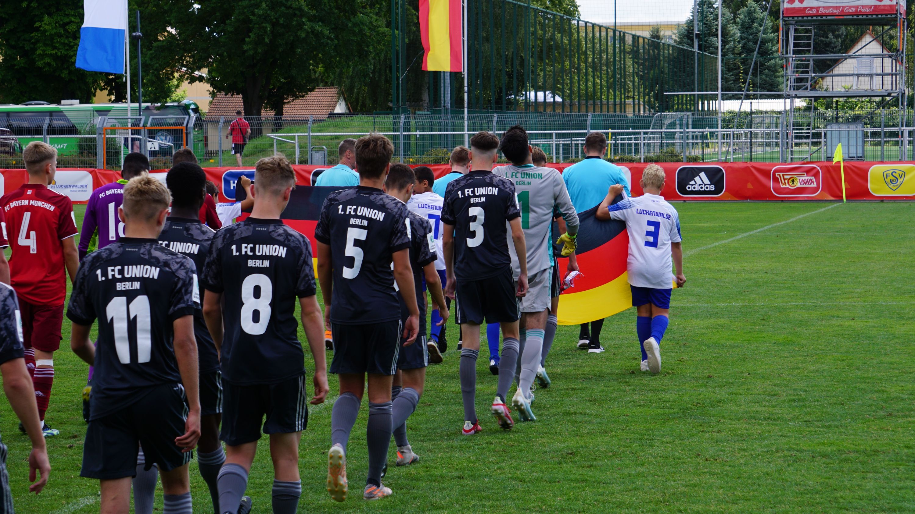 Team lineup of players in soccer jerseys entering a sports field, accompanied by a flag.