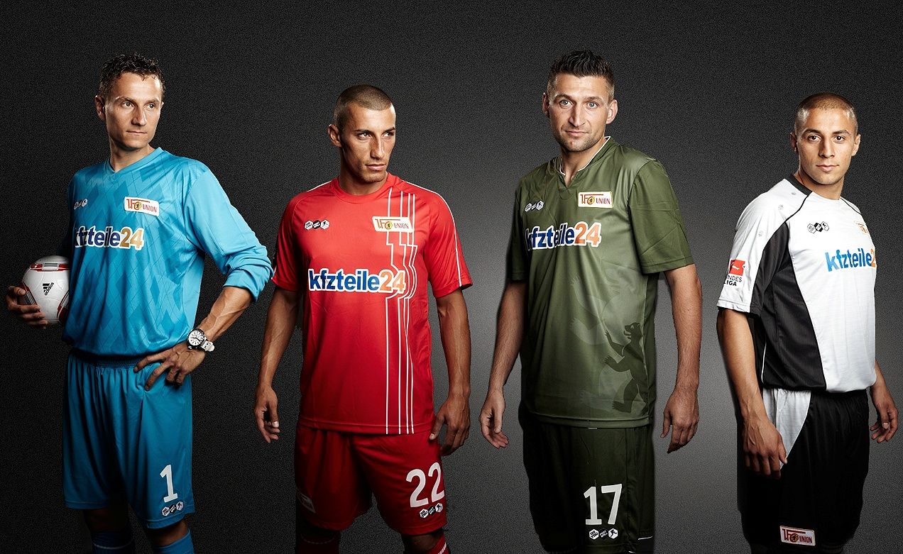 Vier Fußballspieler in verschiedenen Trikots und einer Torwartausstattung posieren vor einem dunklen Hintergrund.