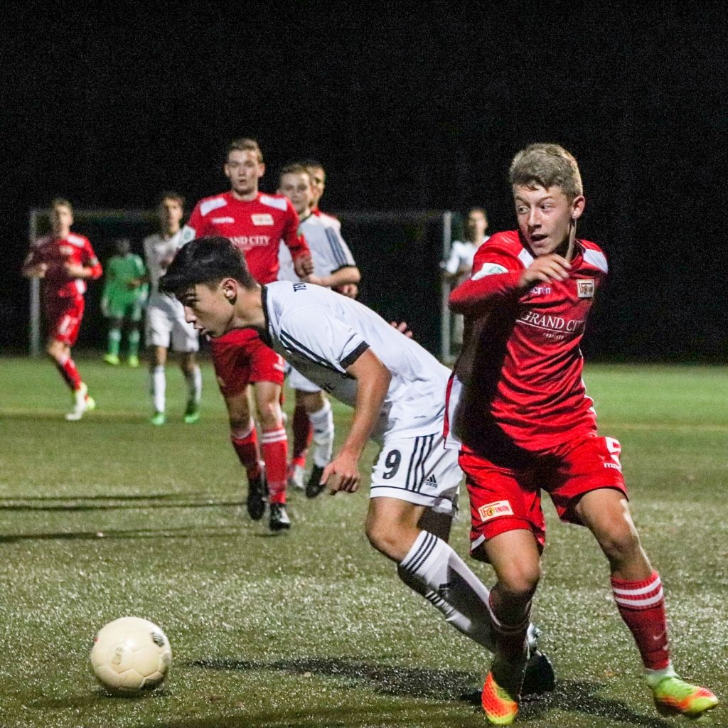 Ein Fußballspiel bei Nacht, zwei Spieler in weißen und roten Trikots verfolgen den Ball auf dem Rasen.