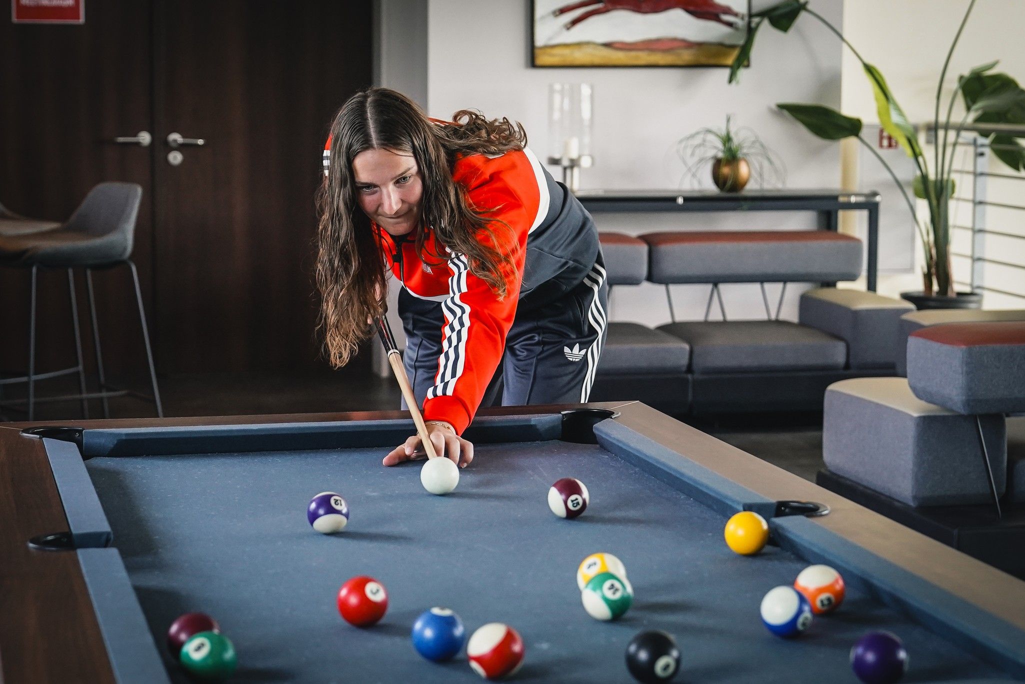 Eine Person mit langen Haaren spielt Billard und konzentriert sich auf den weißen Ball am Tisch mit bunten Kugeln.