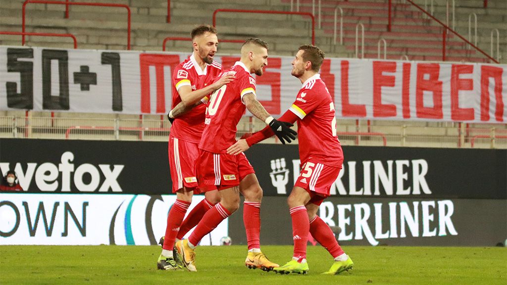 Three football players celebrate a goal in a stadium, with a banner visible in the background.