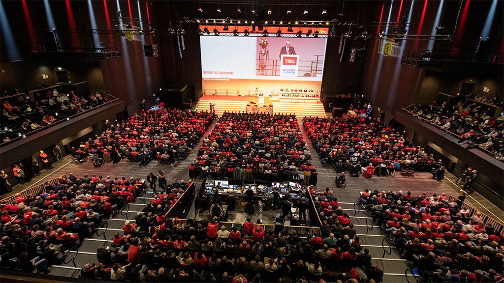 Große Versammlung in einem ausverkauften Veranstaltungsraum mit Menschen in roten Shirts, auf einer Bühne spricht eine Person.