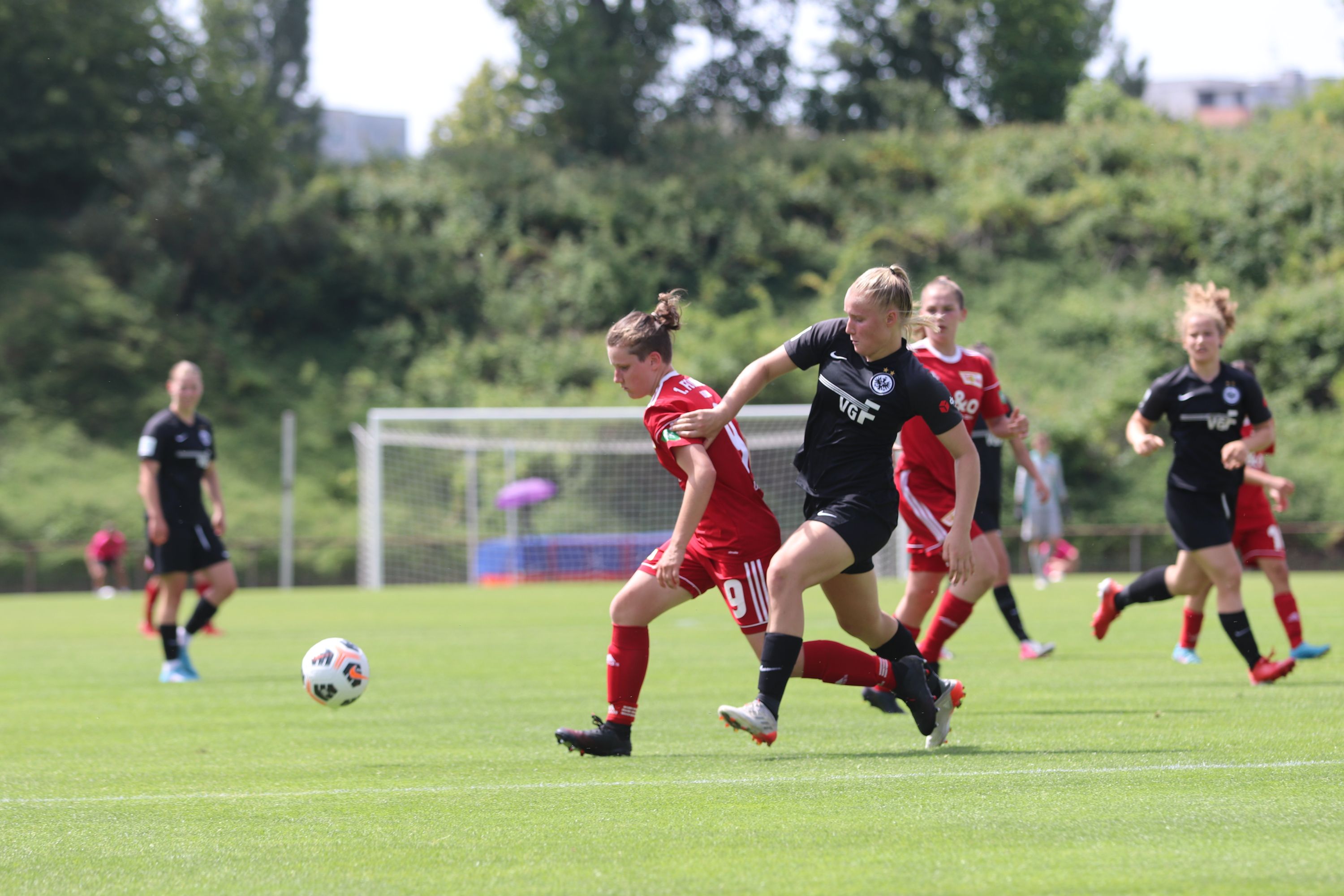 Zwei Fußballspielerinnen im Wettkampf um den Ball auf einem grünen Rasenplatz, im Hintergrund stehen weitere Spielerinnen.