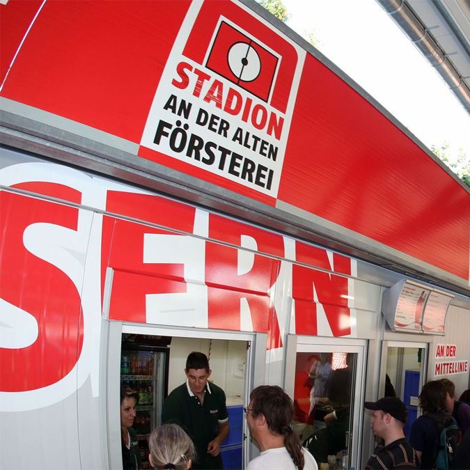 Schilder und Verkaufsstand eines Stadions mit Aufschrift "Stadion an der alten Försterei". Menschen stehen an einem Verkaufsfenster.