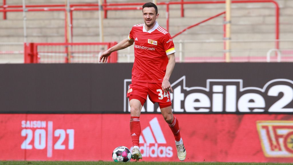 A soccer player in a red jersey dribbles the ball on the field, with a stadium in the background.