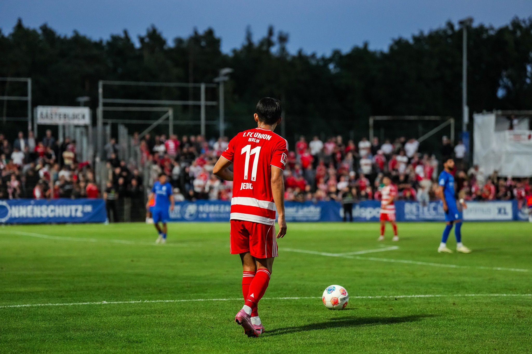 Spieler in roter Trikotausrüstung mit der Nummer 11 steht auf dem Fußballfeld, im Hintergrund Fans und andere Spieler.